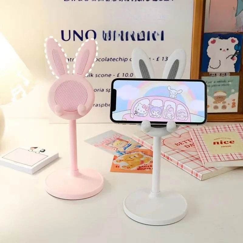 Two phone stands, one pink with bunny ears and one white, on a desk.